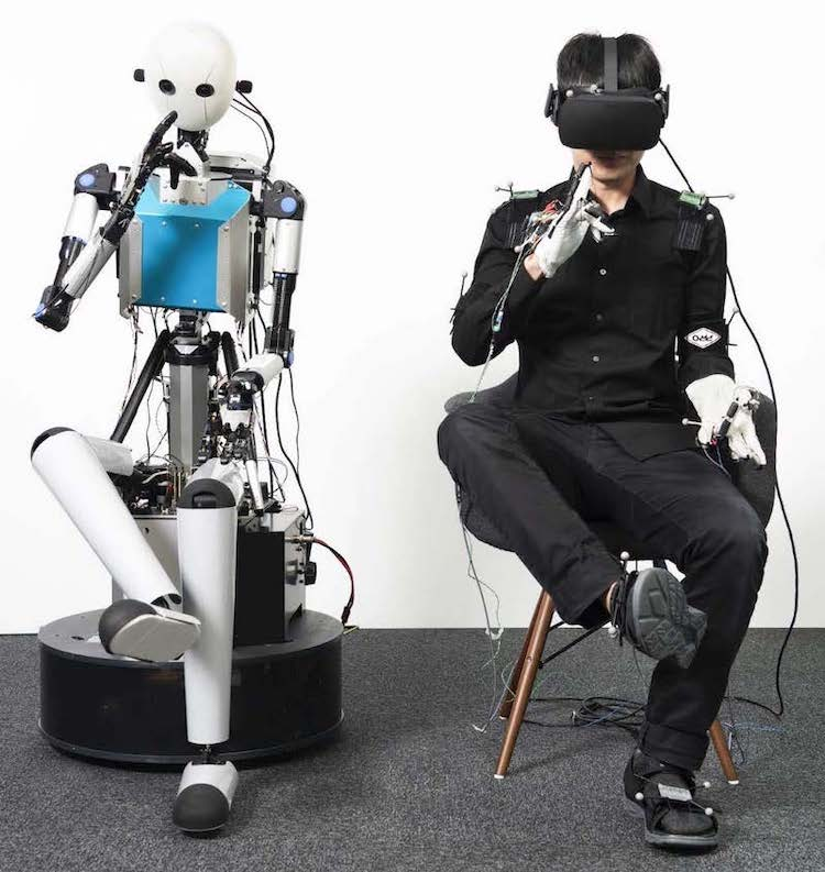 A humanoid robot beside a seated operator wearing a VR headset and haptic gloves